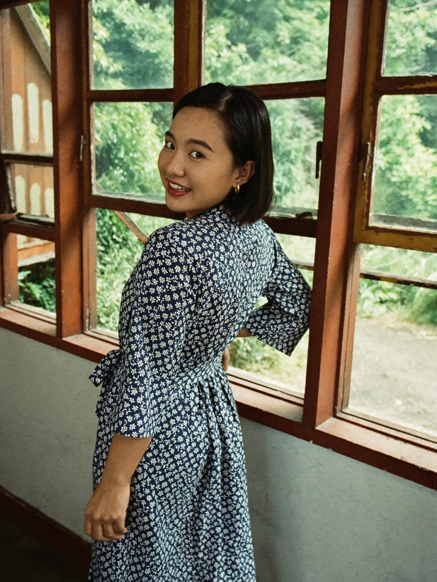 Perempuan dengan terusan navy bermotif bunga menoleh di dekat jendela kayu