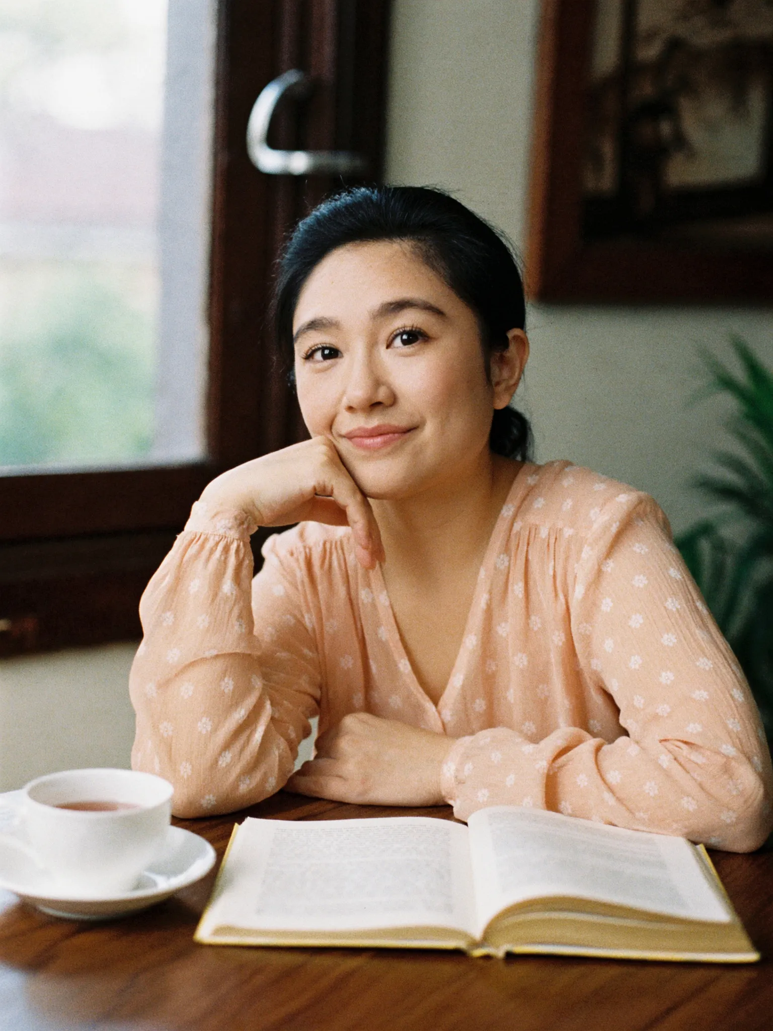 Perempuan dengan blus peach bermotif bunga duduk di meja kayu dengan secangkir teh dan buku