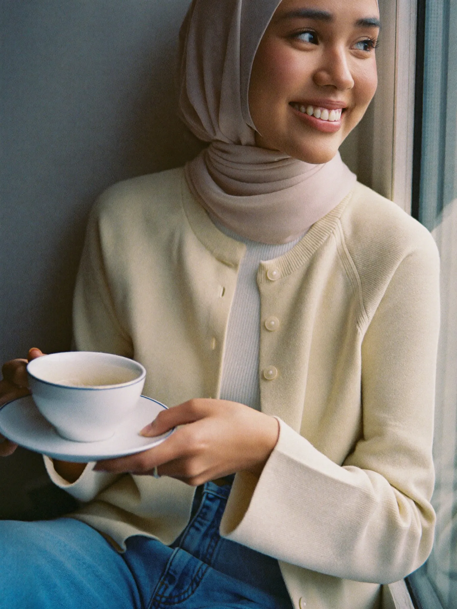 Perempuan berhijab dengan kardigan krem memegang cangkir teh di dekat jendela